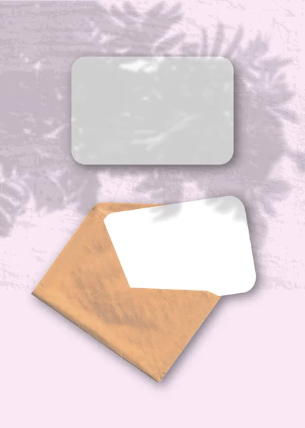 Une enveloppe avec deux feuilles de papier blanc texturé sur le fond rose de la table. Maquette superposée avec les ombres de la plante. La lumière naturelle projette des ombres d'une branche de Rowan — Photo
