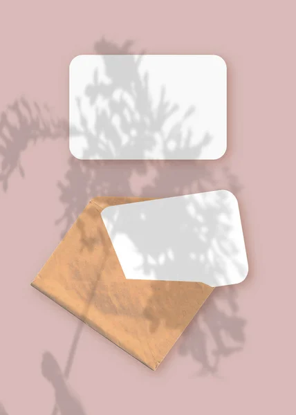 Mockup met een overlay van plantenschaduwen op envelop met twee vellen getextureerd wit papier op een roze tafelachtergrond. Verticale oriëntatie — Stockfoto