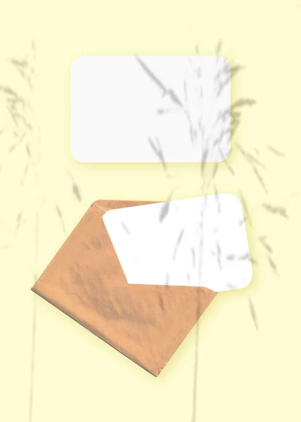 在信封上贴上一层植物阴影的模型，在黄色的桌子背景上贴上两张有纹理的白纸。纵向方向 — 图库照片