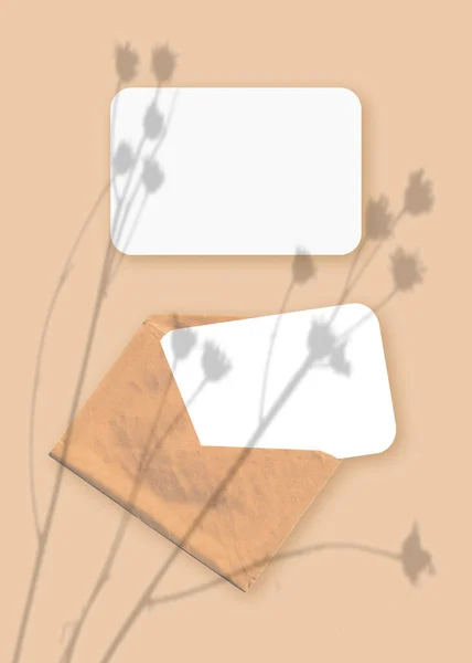 Mockup dengan lapisan bayangan tanaman pada amplop dengan dua lembar kertas putih bertekstur pada latar belakang meja beige. Orientasi vertikal — Stok Foto