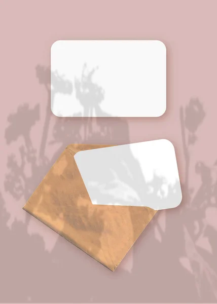Mockup dengan lapisan bayangan tanaman pada amplop dengan dua lembar kertas putih bertekstur pada latar belakang meja merah muda. Orientasi vertikal — Stok Foto