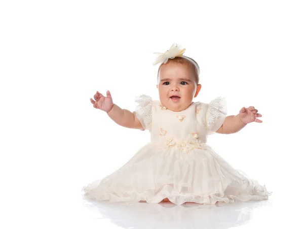 Vacker baby barn i en festlig klänning och ett pannband, sitter på golvet och glatt höjde händerna. skott på en vit bakgrund. — Stockfoto