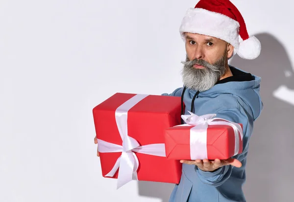 Confident stylish middle aged man in a Santa Claus hat holds out a gift to the camera. — Stock Photo, Image