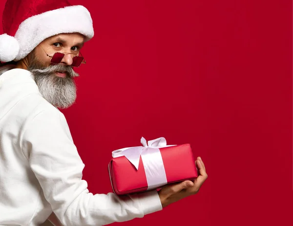 Vieil homme à la mode en chapeau de Père Noël et barbe blanche vous offre un cadeau. sur fond rouge. — Photo