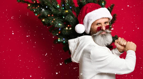 Père Noël senior à la mode debout avec un arbre de Noël dans ses mains sous la neige. — Photo