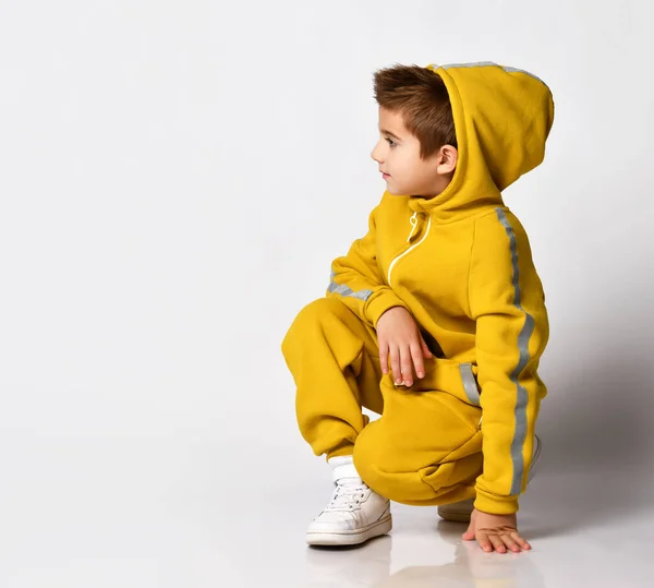 Alegre niño juguetón en un traje de invierno caliente de moda de deportes amarillos sentado sobre un fondo gris. —  Fotos de Stock