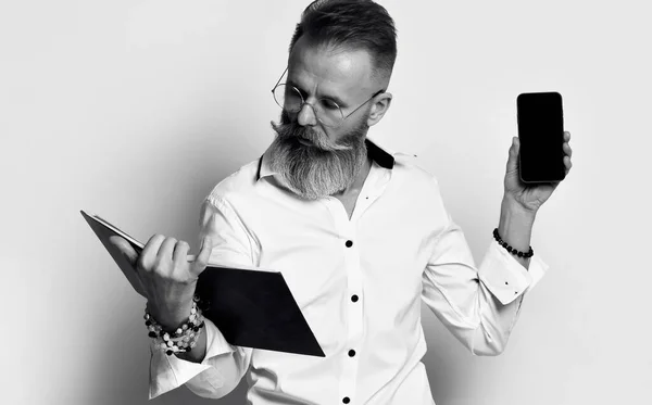 Gri sakallı şık, vahşi bir adam elinde bir kitap ya da günlük tutuyor ve cep telefonunun ekranını gösteriyor. — Stok fotoğraf