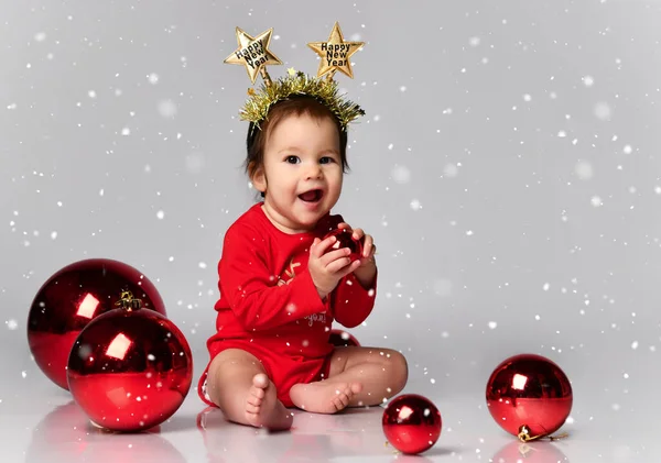 Criança pequena vestindo uma faixa de cabeça com a inscrição Feliz Natal no chão dentro de casa, cercada por bolas de Natal. Primeiro Natal — Fotografia de Stock