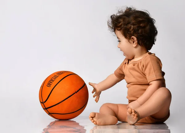 Μικρό αγόρι κάθεται ξυπόλητο και παίζει ένα νέο μπάσκετ σε γκρι φόντο. — Φωτογραφία Αρχείου