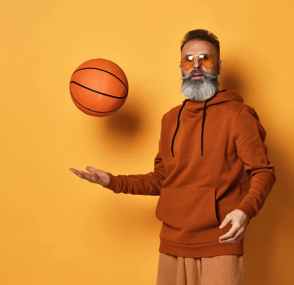 Homem barbudo sério em óculos de sol, com capuz segurando bola cesta — Fotografia de Stock