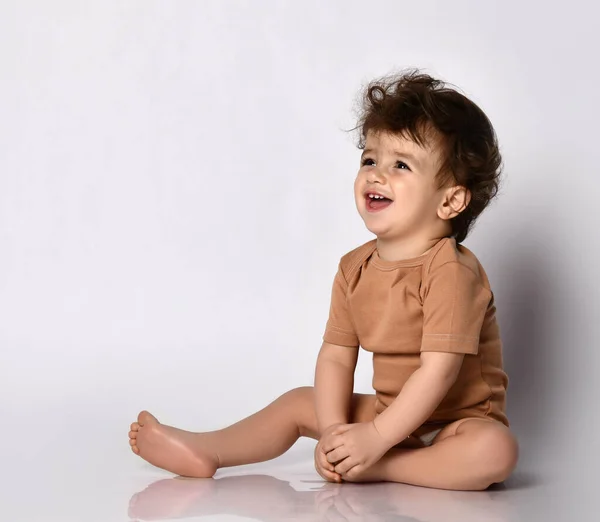 Liten lockig pojke klädd i brun body suit skrattar högt sittande barfota på en grå bakgrund. — Stockfoto