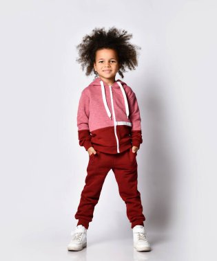 Spor giyimli, kıvırcık saçlı, spor ayakkabılı, spor ayakkabılı, cebinde elleri olan, beyaz arka planda izole edilmiş kameraya poz veren küçük bir Afrikalı Amerikalı kız modelinin stüdyo fotoğrafı..