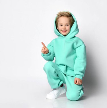 Spor takım elbiseli ve kafasında başlığı olan sarışın bir çocuk gri bir arka planda oturuyor..