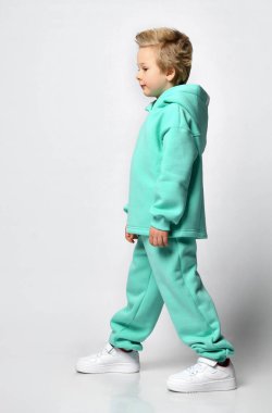 Gri bir arka planda kalın turkuaz takım elbiseli mutlu sarışın çocuk..