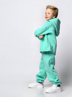 Gri bir arka planda kalın turkuaz takım elbiseli mutlu sarışın çocuk..