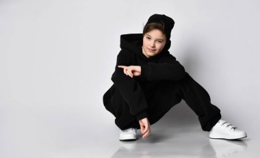 Schoolboy in a stylish black tracksuit and white sneakers posing on a white background. The child looks to the side, sitting near the free space for the text. clipart