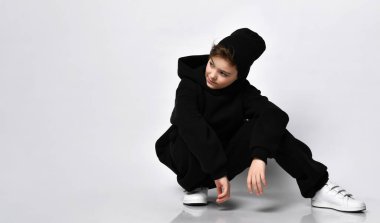 Siyah eşofmanlı ve beyaz arka planda poz veren beyaz spor ayakkabılı bir öğrenci. Çocuk yan tarafa bakıyor, metin için boş alana yakın oturuyor..