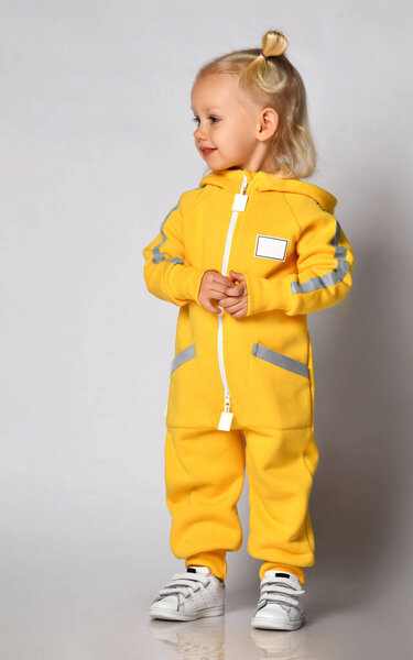 Маленькая симпатичная блондинка в теплом удобном комбинезоне, стоящая напротив серой стены. Счастливого детства и модной детской одежды