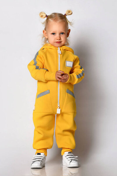 Маленькая симпатичная блондинка в теплом удобном комбинезоне, стоящая напротив серой стены. Счастливого детства и модной детской одежды