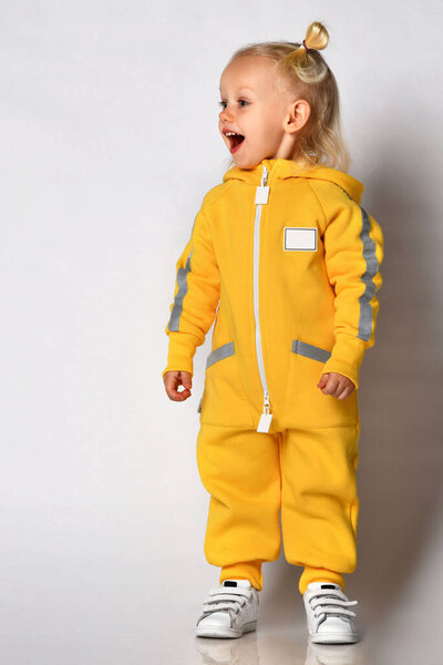 Радостно удивленная маленькая девочка в теплом спортивном комбинезоне на сером фоне.