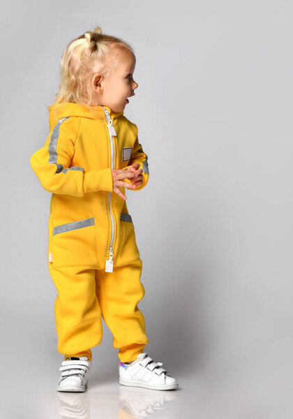 Маленькая симпатичная блондинка в теплом удобном комбинезоне, стоящая напротив серой стены. Счастливого детства и модной детской одежды