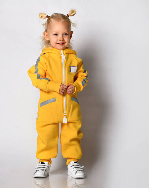 Маленькая симпатичная блондинка в теплом удобном комбинезоне, стоящая напротив серой стены. Счастливого детства и модной детской одежды