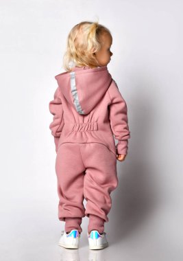 Gri bir duvara karşı duran, sıcak, rahat tulumlu, tatlı sarışın kız. Mutlu çocukluk ve şık bebek kıyafetleri