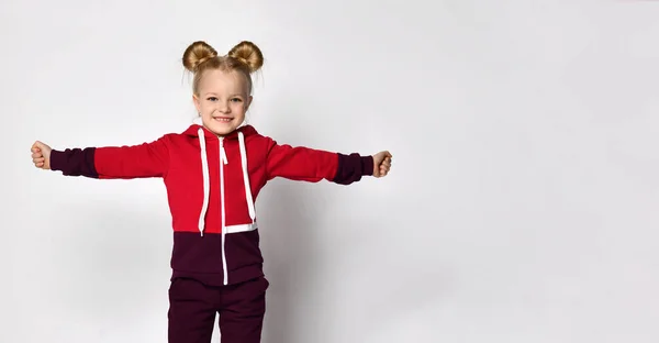 Pequena loira de cabelos cacheados linda menina em um terno vermelho burgundy fica com os braços abertos como asas, jogando um avião ou um pássaro em um fundo de parede cinza. — Fotografia de Stock