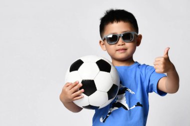 Küçük bir çocuk futbol topuyla kameraya baş parmak işareti yapıyor.