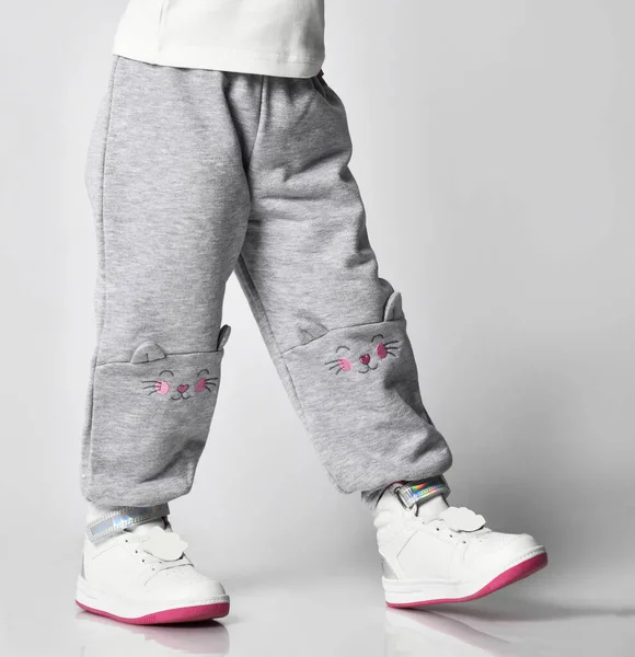Primer plano de un niño en pantalones deportivos grises con un estampado de una cara de gato y zapatillas blancas sobre un fondo gris. Primer plano — Foto de Stock