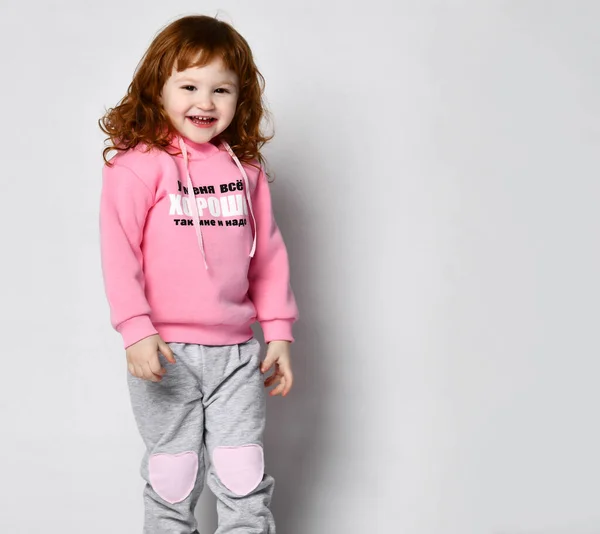 Geïsoleerde afbeelding van een schattige schattige roodharige baby meisje in een roze belettering hoodie poseren in een lege studio met een zelfverzekerde vrolijke glimlach. — Stockfoto
