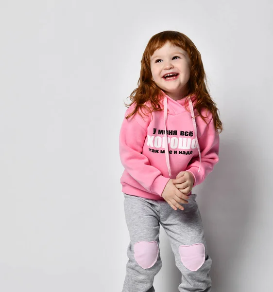 Geïsoleerde afbeelding van een schattige schattige roodharige baby meisje in een roze belettering hoodie poseren in een lege studio met een zelfverzekerde vrolijke glimlach. — Stockfoto