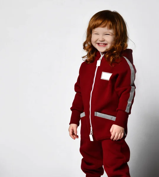 Piccola bella ragazza dai capelli rossi in una tuta rossa ride su uno sfondo grigio muro. Elegante moda casual per bambini. — Foto Stock