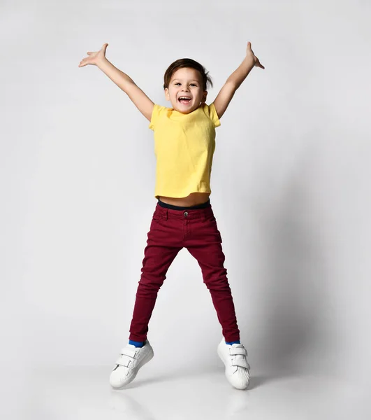 portrait of fashionable preschool little boy in yellow t-shirt, red denim pants, white sneakers, standing isolated on light background