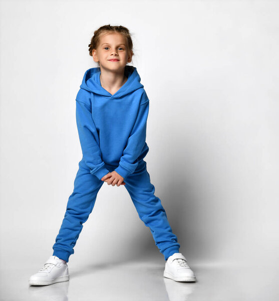 Smiling cute little girl in a blue tracksuit with a hood and trousers, white sneakers, isolated on white. Portrait of a child in cool clothes. Kids fashion, casual wear, comfortable kids wear