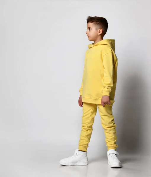 Stijlvolle brunette jongen in een gele sport sweater met een capuchon en broek staat met een capuchon over zijn hoofd, handen in zijn zakken. Straat stijl accessoires trends. Kijk weg.. — Stockfoto