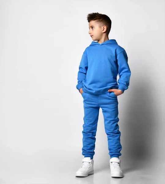 Full-length portret van een jonge knappe man met een stijlvol kapsel in pastel moderne blauwe tinten, met een kap, een sweater, staande, handen in zakken — Stockfoto