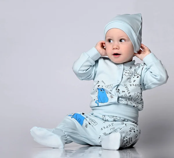 긴 소매가 있는 몸의 아기와, 옷걸이가 있고, 모자가 매칭되어 있고, 옷에 우스꽝 스러운 무늬가 있는, 스튜디오의 바닥에 앉아 있는 아기. — 스톡 사진