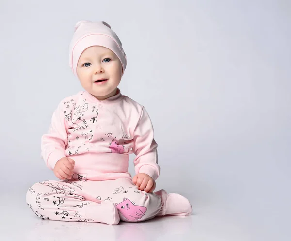 Baby spädbarn i en kropp med långa ärmar och en shianishka, en matchande hatt, med ett roligt barnsligt mönster på sina kläder, sitter på golvet i studion. — Stockfoto