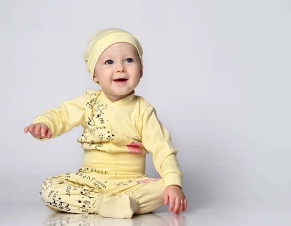 Baby spädbarn i en kropp med långa ärmar och en shianishka, en matchande hatt, med ett roligt barnsligt mönster på sina kläder, sitter på golvet i studion. — Stockfoto