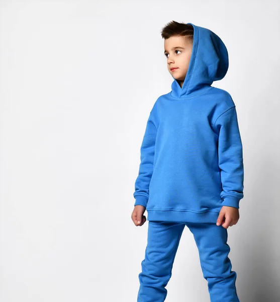 Stílusos fiú, kék sportpulóverben, kapucnival és nadrággal, csuklyával a fején áll, és mutat valahová oldalra. Utcai kiegészítők trendek. Fordulj el!. — Stock Fotó