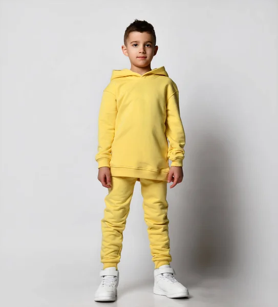 Snygg brunett pojke barn i en sport tröja med en huva och byxor står med en huva över huvudet, händerna i fickorna. — Stockfoto