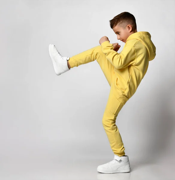 Αγόρι ντυμένο με ένα φωτεινό κίτρινο αθλητικό κοστούμι και λευκά αθλητικά παπούτσια διασκεδάζει σε γκρι φόντο. — Φωτογραφία Αρχείου