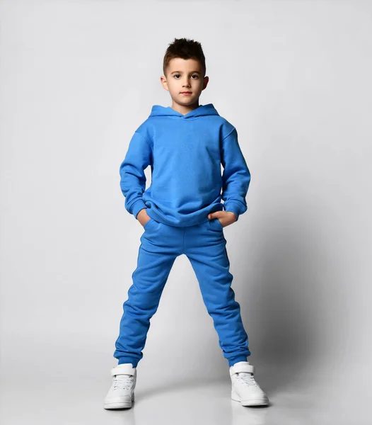 Pastel mavi tonlarda, kapüşonlu, kazaklı, ayakta duran, elleri cepli, şık bir saç kesimi olan genç bir adamın tam boy portresi.