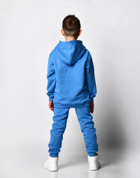 Retrato completo de um menino com um corte de cabelo elegante em tons pastel modernos de azul com um capuz e calças, de pé com as costas para a câmera sobre fundo branco branco. — Fotografia de Stock