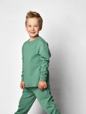 Yeşil renkli rahat bir takım elbise giymiş neşeli küçük çocuk, yarım yamalak duruyor ve hafif bir arkaplanda kameraya neşeyle bakıyor.