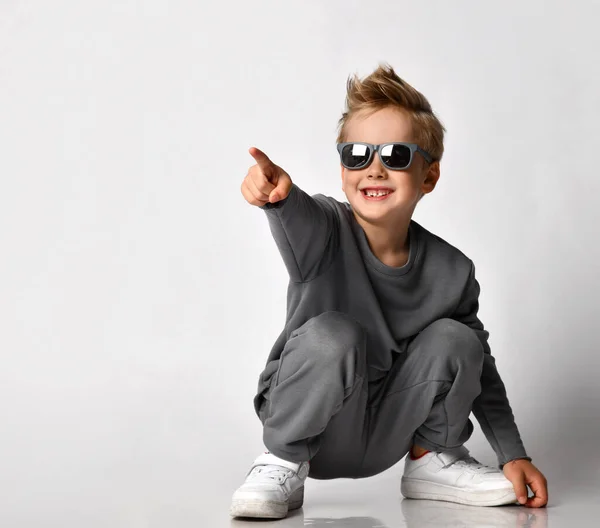 Little fashion boy squatting pointing finger forward studio shot — Stock fotografie