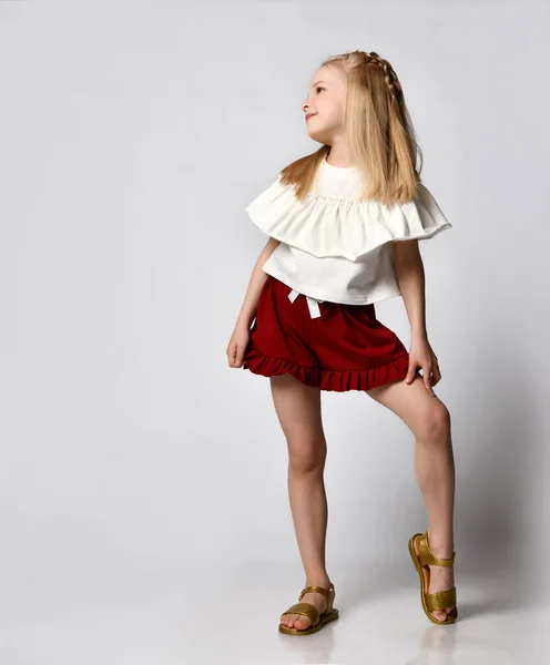 Cute smiling little girl in a white blouse with ruffles, burgundy shorts and sandals, put her leg to the side — Stock Photo, Image