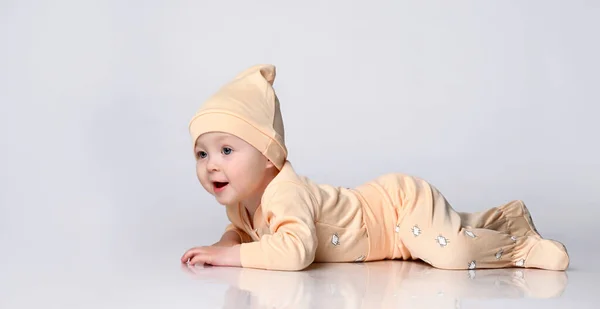 Adorable laughing little kid in a yellow cotton jumpsuit and a hat is lying on the floor and trying to crawl. on white background. — Stock Photo, Image