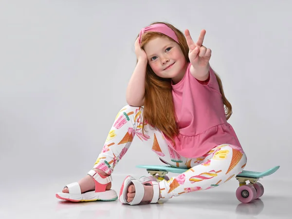 Glimlachend klein roodharig meisje zittend op skateboard gebaar van overwinning — Stockfoto